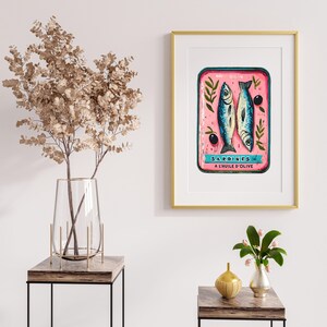 May include: A vintage-style illustration of a can of sardines in olive oil. The can is pink with blue and white lettering that reads "SARDINES A L'HUILE D'OLIVE". The illustration is framed in a gold frame.