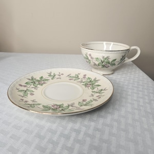 May include: A cream-colored teacup and saucer set. The cup and saucer feature a floral design with green leaves and small pink flowers. Both pieces have a gold trim. The set is placed on a light-colored surface.