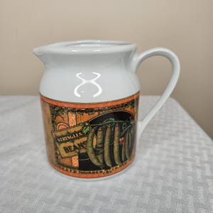 Op de afbeelding: Witte keramische creamer met een handvat en tuit. De creamer heeft een decoratieve band met een vintage illustratie van sperziebonen. De tekst "STRINGLESS BEANS" is zichtbaar op het ontwerp. De creamer heeft een klassieke, boerderij-esthetiek.