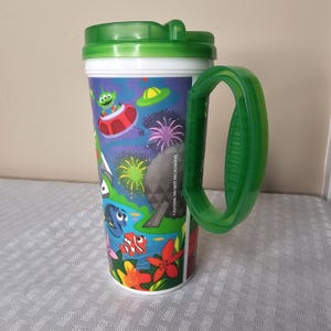 May include: A green-handled travel mug with a green lid and a white rim. The mug features a colorful cartoon design with aliens, spaceships, and underwater scenes. The text "CAUTION: DO NOT MICROWAVE" is printed on the side.