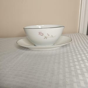 Könnte beinhalten: Ein weißes Porzellan-Sauciere- und Untertassen-Set. Die Sauciere hat einen schwarzen Rand und ein zartes rosa Rosendesign. Die Untertasse hat ebenfalls einen schwarzen Rand. Das Set steht auf einer weiß gemusterten Tischdecke.