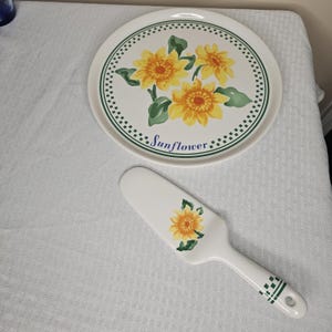May include: White ceramic serving plate and pie server with a sunflower design. The plate has a green dotted border and the word "Sunflower" in blue script. The pie server has a matching sunflower design and green accents.