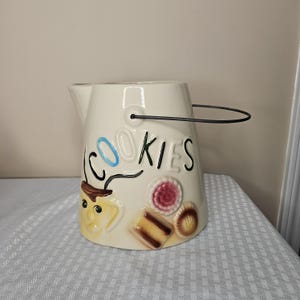May include: Cream ceramic cookie jar with a black handle. The word "COOKIES" is written on the side in blue and black lettering. The jar is decorated with embossed cookie designs in various colours.