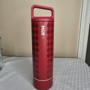 K&ouml;nnte beinhalten: Eine rote, isolierte Wasserflasche mit Griff. Die Flasche hat ein Rautenmuster und den Markennamen "Miir+" in Wei&szlig;. Der Boden der Flasche hat ein goldenes Band.