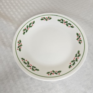 Peut inclure: Assiette en céramique blanche avec une bordure verte et un motif de baies de houx. L'assiette présente des feuilles de houx vertes et des baies rouges autour du bord, créant un thème de vacances festif. L'assiette est ronde et a un design simple et élégant.