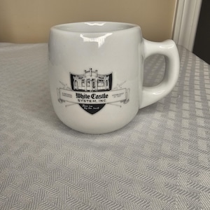 May include: White ceramic mug with a rounded base and a large handle. The mug features a black and white graphic of a building with the text "White Castle System, Inc." and "Established in 1921". The mug is on a white patterned surface.