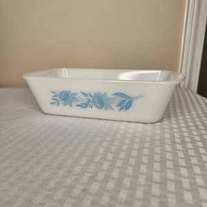 May include: A rectangular, white glass baking dish with a floral design in light blue. The flowers and leaves are arranged in a horizontal pattern across the front of the dish. The dish has a simple, classic design.