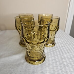 Könnte beinhalten: Sechs bernsteinfarbene Trinkgläser. Jedes Glas hat ein strukturiertes, facettiertes Design im unteren Bereich und einen glatten, zylindrischen oberen Abschnitt. Die Gläser stehen auf einer weißen Tischdecke.