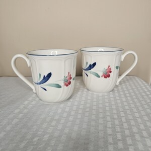 Puede incluir: Dos tazas de cerámica blanca con un diseño floral. Cada taza presenta un borde y un asa azules, con un patrón floral pintado en azul, rojo y verde. Las tazas están sobre un mantel blanco estampado.