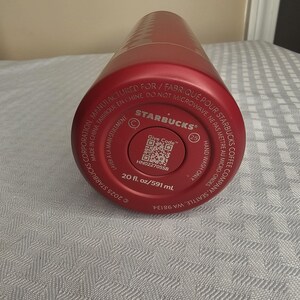 K&ouml;nnte beinhalten: Ein roter Starbucks-Becher mit einem strukturierten Muster. Der Boden des Bechers zeigt das Starbucks-Logo, einen QR-Code und Text, der eine Kapazit&auml;t von 591 ml angibt. Der Text weist auch darauf hin, dass der Becher nur von Hand gewaschen werden darf.