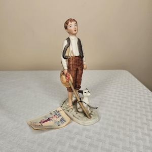 Puede incluir: Una figura de porcelana de un niño con cabello castaño, que lleva una camisa blanca, pantalones marrones y un chaleco negro. Sostiene un sombrero y un palo de madera, con un perro pequeño a sus pies. Una etiqueta con texto adjunta.