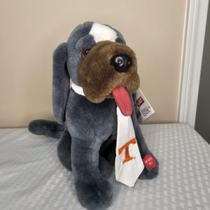 May include: A plush dog toy in shades of gray, brown, and white, with a red tongue sticking out. It wears a white scarf with an orange "T" logo. The dog has a white patch on its head and a tag attached.