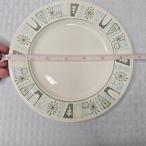 Op de afbeelding: Crème-witte keramische plaat met een decoratieve rand met geometrische vormen en sterren in zwart, lichtblauw en groen. De plaat heeft een diameter van ongeveer 18 cm.