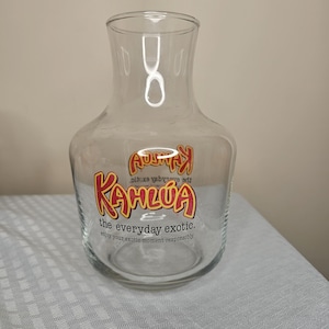 Può includere: Una caraffa di vetro trasparente con una base larga e un collo stretto. La caraffa presenta il logo "Kahlúa" in rosso e giallo, con il testo "the everyday exotic" e "enjoy your exotic moment responsibly."