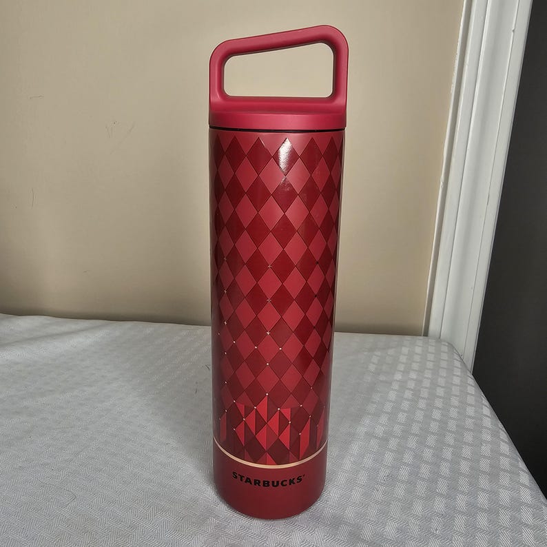 K&ouml;nnte beinhalten: Eine rote Starbucks-Trinkflasche mit Griff. Die Flasche hat ein Rautenmuster und ein goldenes Band am unteren Rand. Das Starbucks-Logo ist auf der Unterseite der Flasche zu sehen. Die Flasche ist etwa 23 cm hoch.