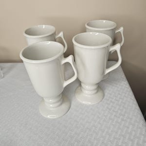 May include: Four white ceramic mugs with handles. The mugs have a pedestal base and a rounded top. The mugs are arranged on a white patterned surface. The mugs are a neutral color and could be used for coffee or tea.