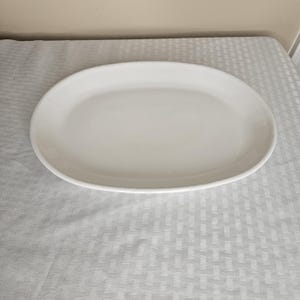 Puede incluir: Una fuente ovalada blanca para servir. La fuente de cerámica tiene una superficie lisa y bordes ligeramente elevados. Está vacía y descansa sobre un mantel blanco. Adecuada para servir alimentos.