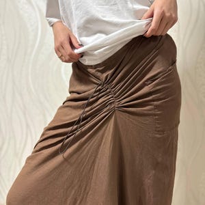 May include: A person wearing a white long-sleeve shirt and a brown skirt. The skirt has a drawstring waist and a gathered design. The skirt is a solid brown color and appears to be made of a soft fabric.