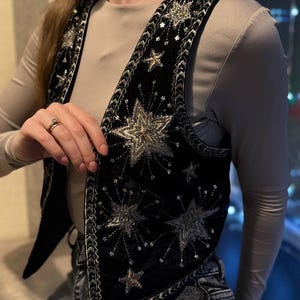 K&ouml;nnte beinhalten: Schwarze Samtweste mit silbernen Sternen und Pailletten verziert. Die Weste hat einen detaillierten silbernen Besatz an den Kanten und um die Sterndesigns. Ein stilvolles Kleidungsst&uuml;ck zum &Uuml;berziehen, das jedem Outfit einen Hauch von himmlischem Flair verleiht.