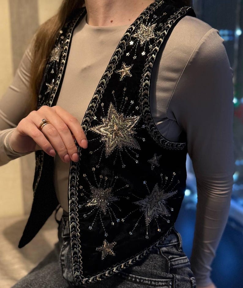 K&ouml;nnte beinhalten: Schwarze Samtweste mit silbernen Sternen und Pailletten verziert. Die Weste hat einen detaillierten silbernen Besatz an den Kanten und um die Sterndesigns. Ein stilvolles Kleidungsst&uuml;ck zum &Uuml;berziehen, das jedem Outfit einen Hauch von himmlischem Flair verleiht.