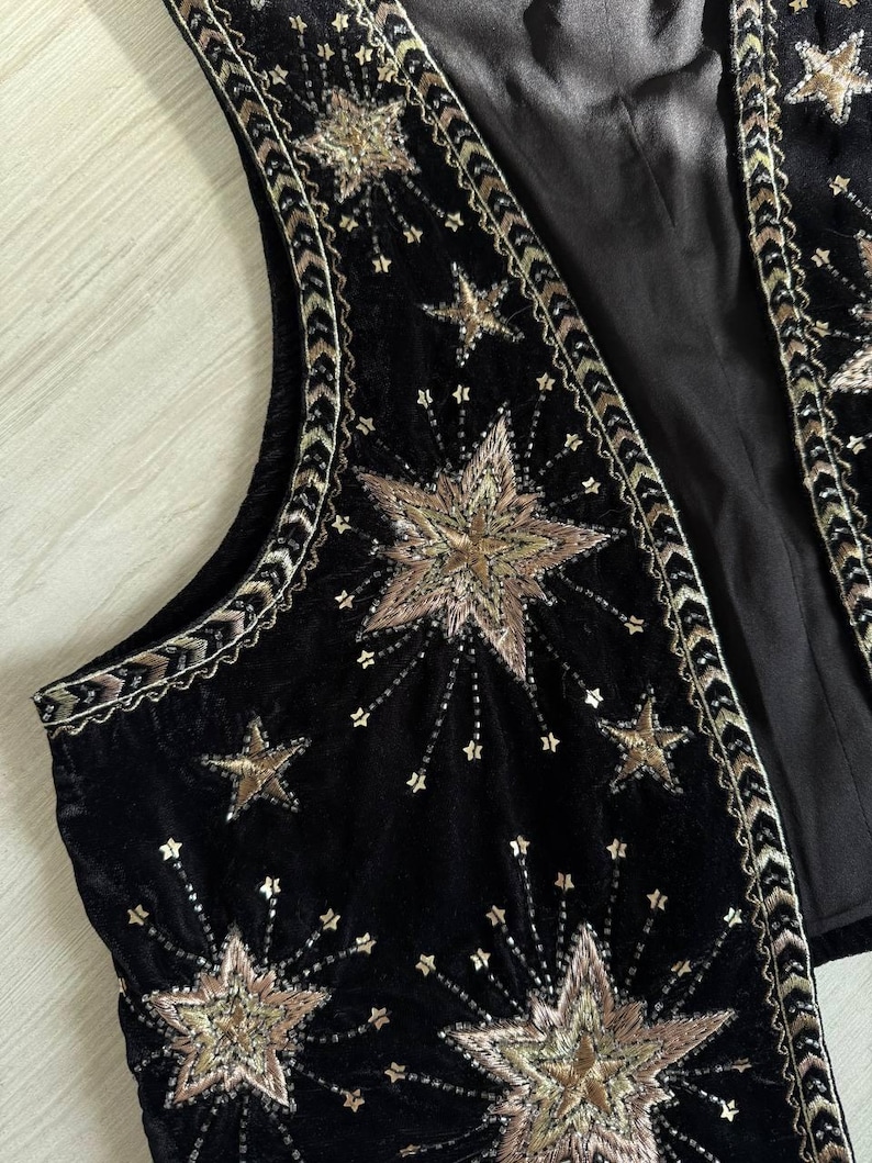 K&ouml;nnte beinhalten: Schwarze Samtweste mit goldener Sternenstickerei. Die Weste hat ein detailliertes Design aus gestickten Sternen in verschiedenen Gr&ouml;&szlig;en mit goldfarbenen Verzierungen an den Kanten. Die Weste hat ein Satinfutter und einen klassischen, &auml;rmellosen Schnitt.