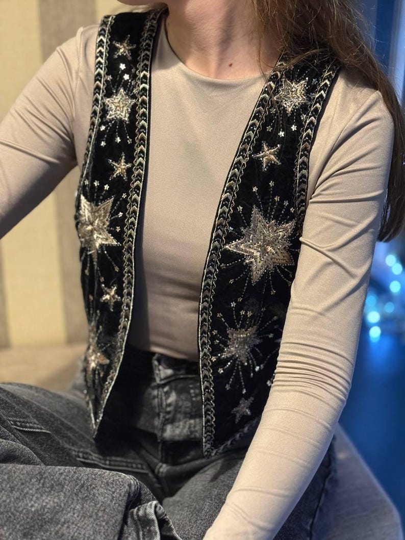 K&ouml;nnte beinhalten: Schwarze Samtweste mit silbernen Sternen und Pailletten. Die Weste hat ein detailliertes Design mit Sternen unterschiedlicher Gr&ouml;&szlig;e und silbernen Verzierungen an den Kanten. Diese Weste ist ein Statement-Piece, ideal f&uuml;r einen Hauch von Glamour.
