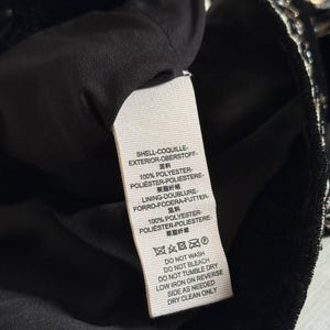 K&ouml;nnte beinhalten: Nahaufnahme eines schwarzen Kleidungsst&uuml;cks mit einem angebrachten Etikett. Das Etikett zeigt Text in mehreren Sprachen, darunter Deutsch, mit Angaben zur Stoffzusammensetzung und Pflegehinweisen. Das Kleidungsst&uuml;ck scheint aus einem weichen, dunklen Material zu bestehen.