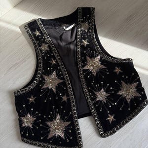 K&ouml;nnte beinhalten: Schwarze Samtweste mit gestickten goldenen und silbernen Sternen. Die Weste hat eine dekorative Bord&uuml;re an den Kanten und Armausschnitten. Ein stilvolles, &auml;rmelloses Kleidungsst&uuml;ck zum &Uuml;berziehen.