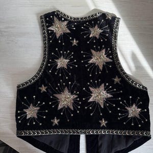 K&ouml;nnte beinhalten: Schwarze Samtweste mit gestickten Sternen und Sternenmustern in Gold und Silber. Die Weste hat eine dekorative Bord&uuml;re und eine taillierte Passform. Ein einzigartiges und stilvolles Kleidungsst&uuml;ck.