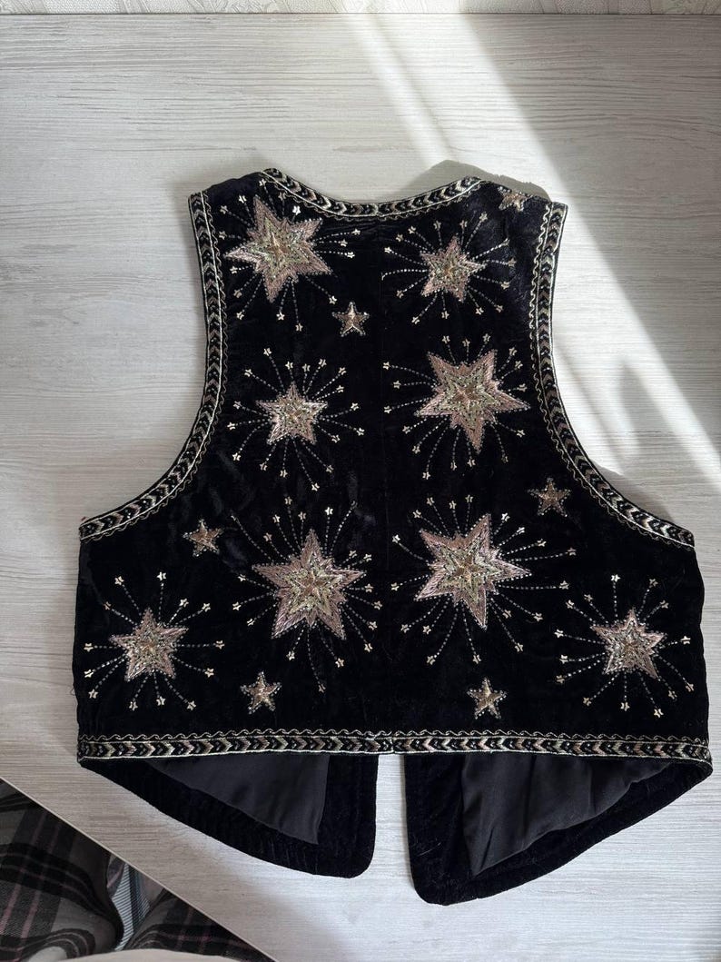 K&ouml;nnte beinhalten: Schwarze Samtweste mit gestickten Sternen und Sternenmustern in Gold und Silber. Die Weste hat eine dekorative Bord&uuml;re und eine taillierte Passform. Ein einzigartiges und stilvolles Kleidungsst&uuml;ck.