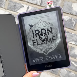 May include: A Kindle e-reader in a protective case, showing the cover of "Iron Flame" by Rebecca Yarros. The book cover has a circular design with the title and author's name. A bookmark with glasses is at the top. The e-reader is held in a hand.