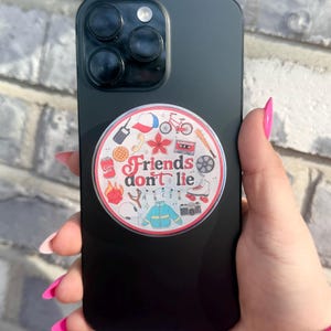 May include: A black smartphone with a circular sticker featuring various cartoon illustrations and the text "Friends don't lie." The sticker includes images of a waffle, a bicycle, and a cassette tape.