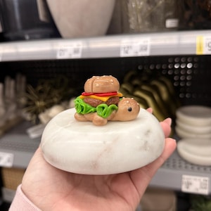May include: A whimsical, handmade turtle figurine with a miniature burger on its back. The burger features a bun, patty, cheese, tomato, and lettuce. The turtle is brown with a white marble base.