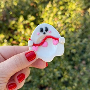 Pu&ograve; includere: Una spilla a forma di fantasma bianco con occhi neri e una bocca piccola. Una linea rossa ondulata si estende attraverso il fantasma, con accenti a forma di cuore rosa. La spilla &egrave; tenuta contro uno sfondo verde sfocato.