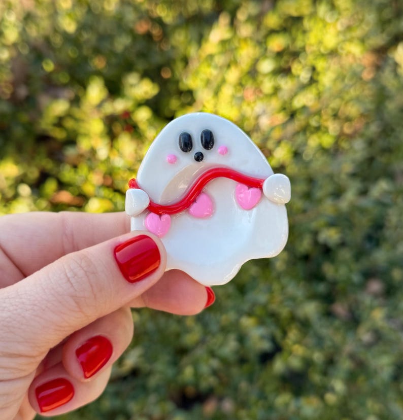Pu&ograve; includere: Una spilla a forma di fantasma bianco con occhi neri e una bocca piccola. Una linea rossa ondulata si estende attraverso il fantasma, con accenti a forma di cuore rosa. La spilla &egrave; tenuta contro uno sfondo verde sfocato.
