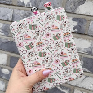 May include: A patterned book cover featuring ghosts, books, flowers, and gifts. Two ghost-shaped bookmarks with glasses are inserted at the top. The design is set against a grey brick wall.