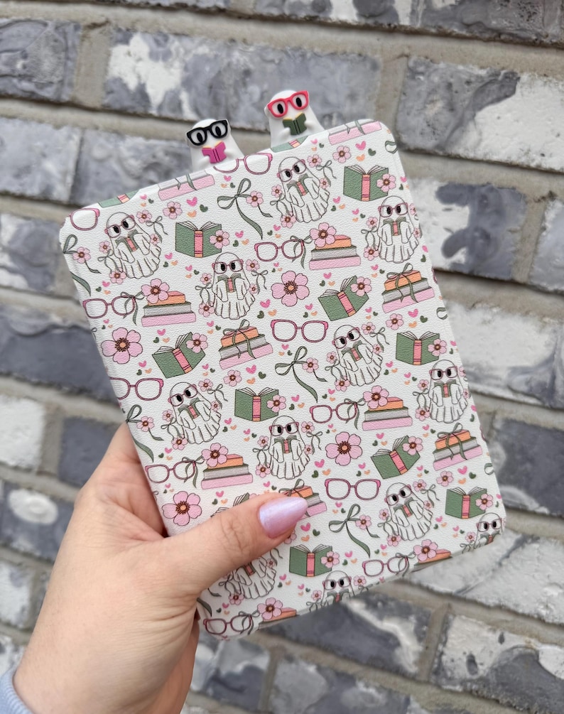 May include: A patterned book cover featuring ghosts, books, flowers, and gifts. Two ghost-shaped bookmarks with glasses are inserted at the top. The design is set against a grey brick wall.