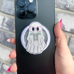 May include: Black smartphone with a pop socket featuring a cartoon ghost wearing pink glasses and reading a book. The ghost is white with a flowing sheet design. The pop socket is set against a light gray background.