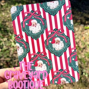 Puede incluir: Un artículo rectangular con temática navideña con un fondo a rayas rojas y blancas. El diseño presenta medallones verdes repetidos con ilustraciones de Papá Noel y lazos rosas. El texto "THE GRAVEYARD BOOTIQUE" es visible.