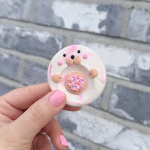 May include: A handmade, circular clay pin featuring a seal design. The pin has a white base with pink marbling, a seal face, and a small cookie in the center. The cookie is pink with colorful sprinkles. The seal has tan paws and a tan nose.