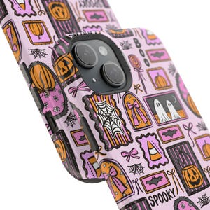 May include: A pink phone case with a Halloween-themed pattern. The design includes pumpkins, ghosts, spiderwebs, bows, and the word "Spooky". The case has a black border and a camera cutout.