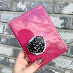 May include: A bright pink phone case with a black and white heart-shaped pop socket that reads "HOT GIRLS READ". The case is held in front of a brick wall.