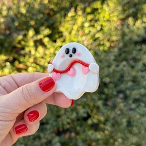 Pu&ograve; includere: Una spilla bianca a forma di fantasma con occhi neri, bocca rossa e una linea ondulata rossa con cuori rosa. La spilla &egrave; tenuta su uno sfondo verde sfocato. Un accessorio affascinante e stravagante.