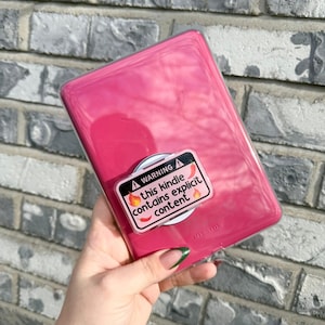 May include: A pink Kindle case with a clear pop-socket featuring the text "WARNING this kindle contains explicit content" and two chili pepper graphics. The case is held in front of a gray brick wall.
