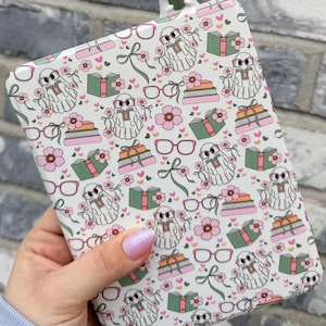 May include: A white e-reader case featuring a pattern of ghosts in glasses, books, flowers, and bows. A matching bookmark with a ghost wearing glasses is at the top. The case is held in a hand with pink nail polish.
