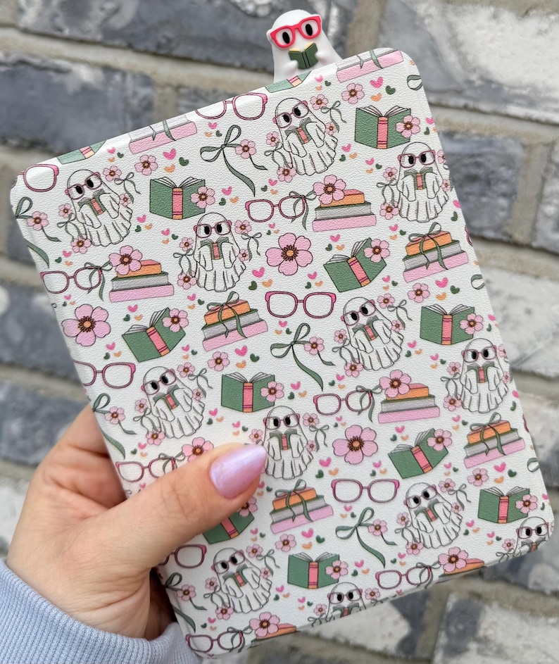 May include: A white e-reader case featuring a pattern of ghosts in glasses, books, flowers, and bows. A matching bookmark with a ghost wearing glasses is at the top. The case is held in a hand with pink nail polish.