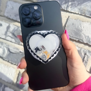 Puede incluir: Smartphone negro con un soporte para teléfono en forma de corazón. El agarre tiene un centro blanco y un borde negro festoneado. Dentro del corazón hay pequeños amuletos de colores. El teléfono se sostiene contra una pared de ladrillos.