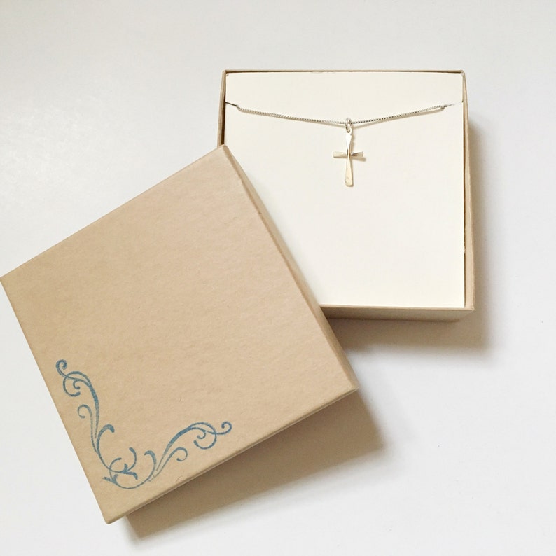 Puede incluir: Un collar con un colgante de cruz de plata en una caja marr&oacute;n con un dise&ntilde;o floral azul. La caja est&aacute; abierta y el collar est&aacute; dentro.