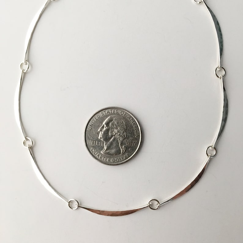 May include: A silver necklace with a hammered finish and a curved design. The necklace is about the size of a quarter.