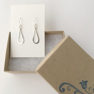 May include: A pair of silver teardrop shaped earrings with a hammered texture, displayed on a white card and in a brown box.